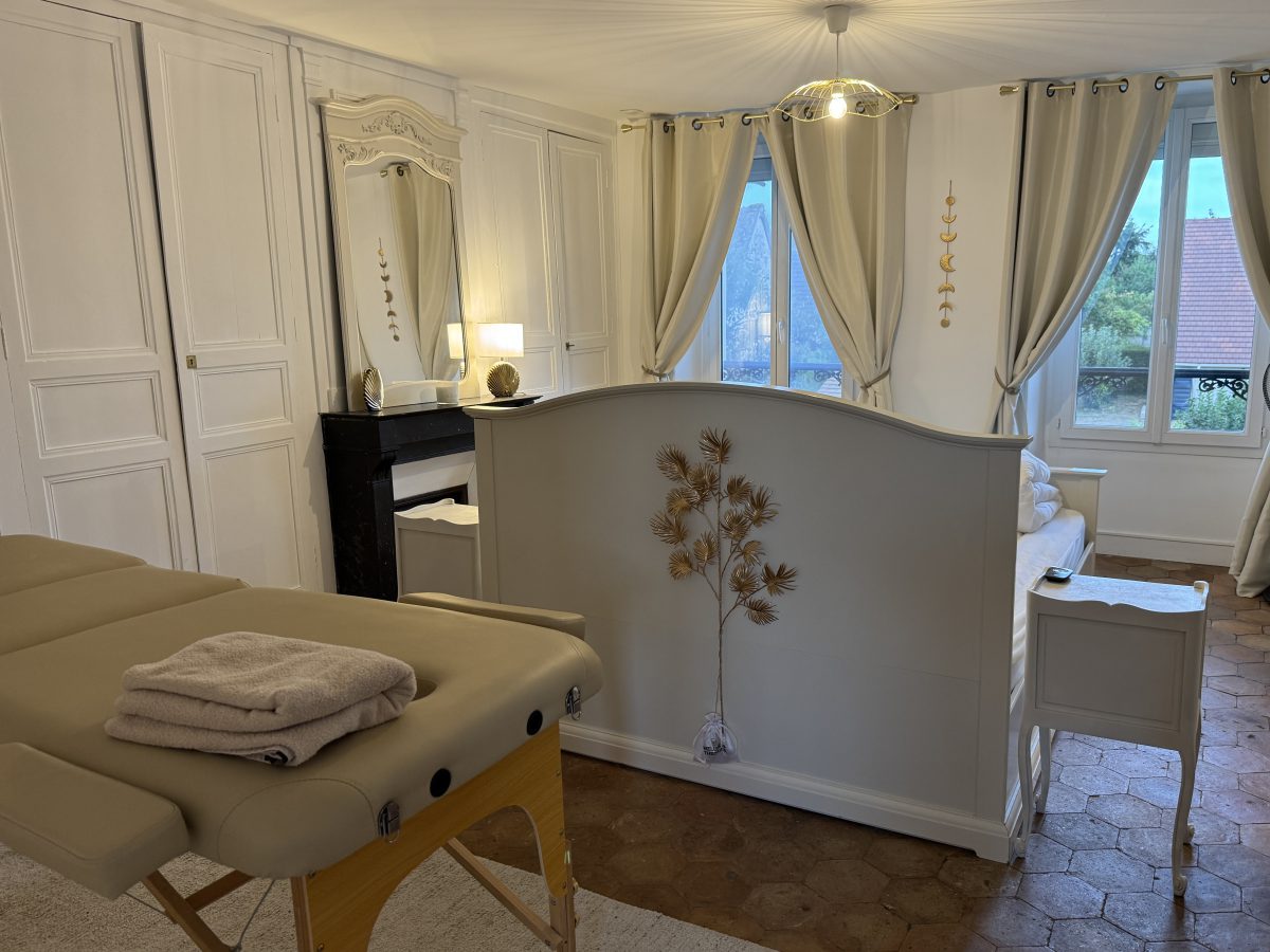 La chambre après travaux : boiseries blanches, tommettes hexagonales, lit derrière une cloison basse, et une table de massage au premier plan